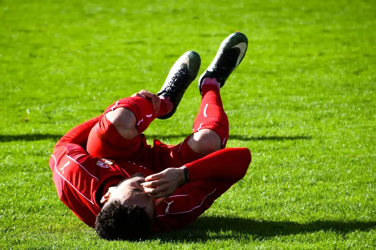 Sportiv în echipament roșu stă întins pe iarbă, ținându-se de față după un disconfort