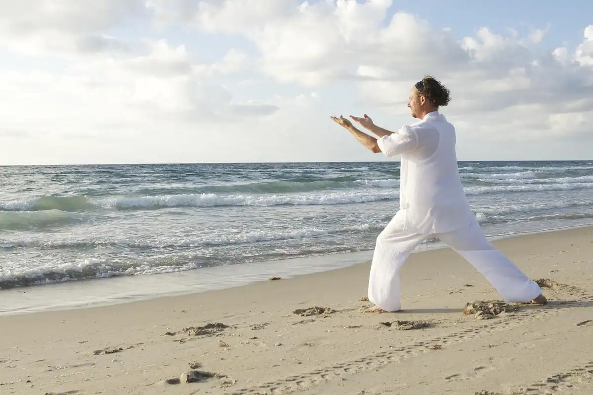 Bărbat în haine albe face mișcări lente de tai chi pe plajă, lângă valuri