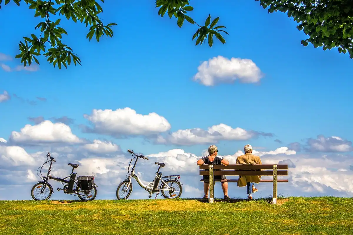 Doi seniori stau pe o bancă, într-un parc, lângă două biciclete electrice